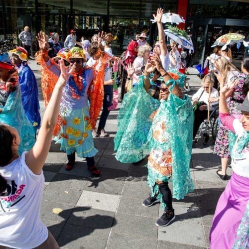 A big group of people throw their hands up in dance wearing brightly coloured carnival costumes - led by charlene low our workshop leader