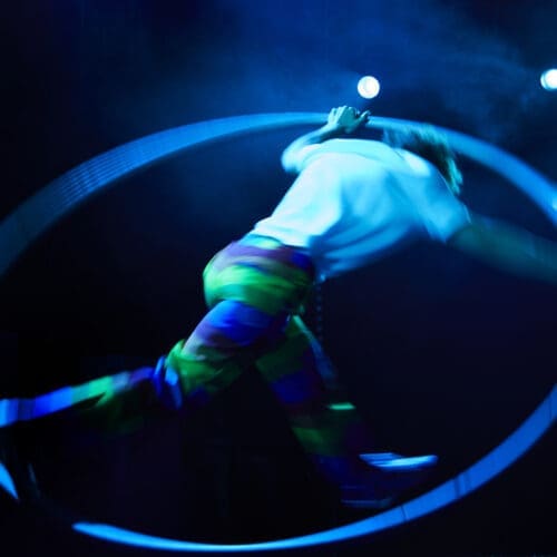 A circus artist spins around inside a cyr wheel under a blue stage spotlight