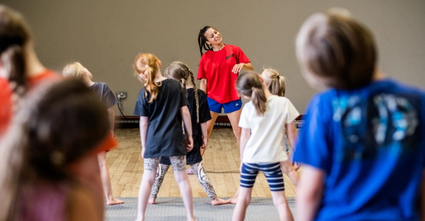 children taking part in a circus summer school with a teacher at the front guiding them in a stretching routine.