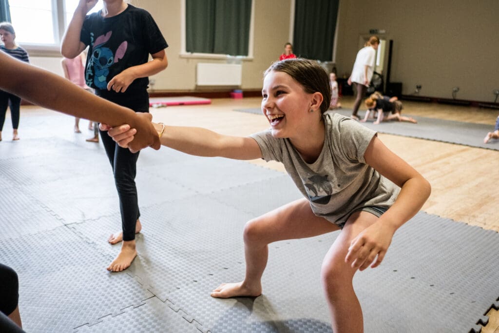 a young girl holds the hand of another and leans back, laughing. she is taking part in a circus summer school in stoke on trent