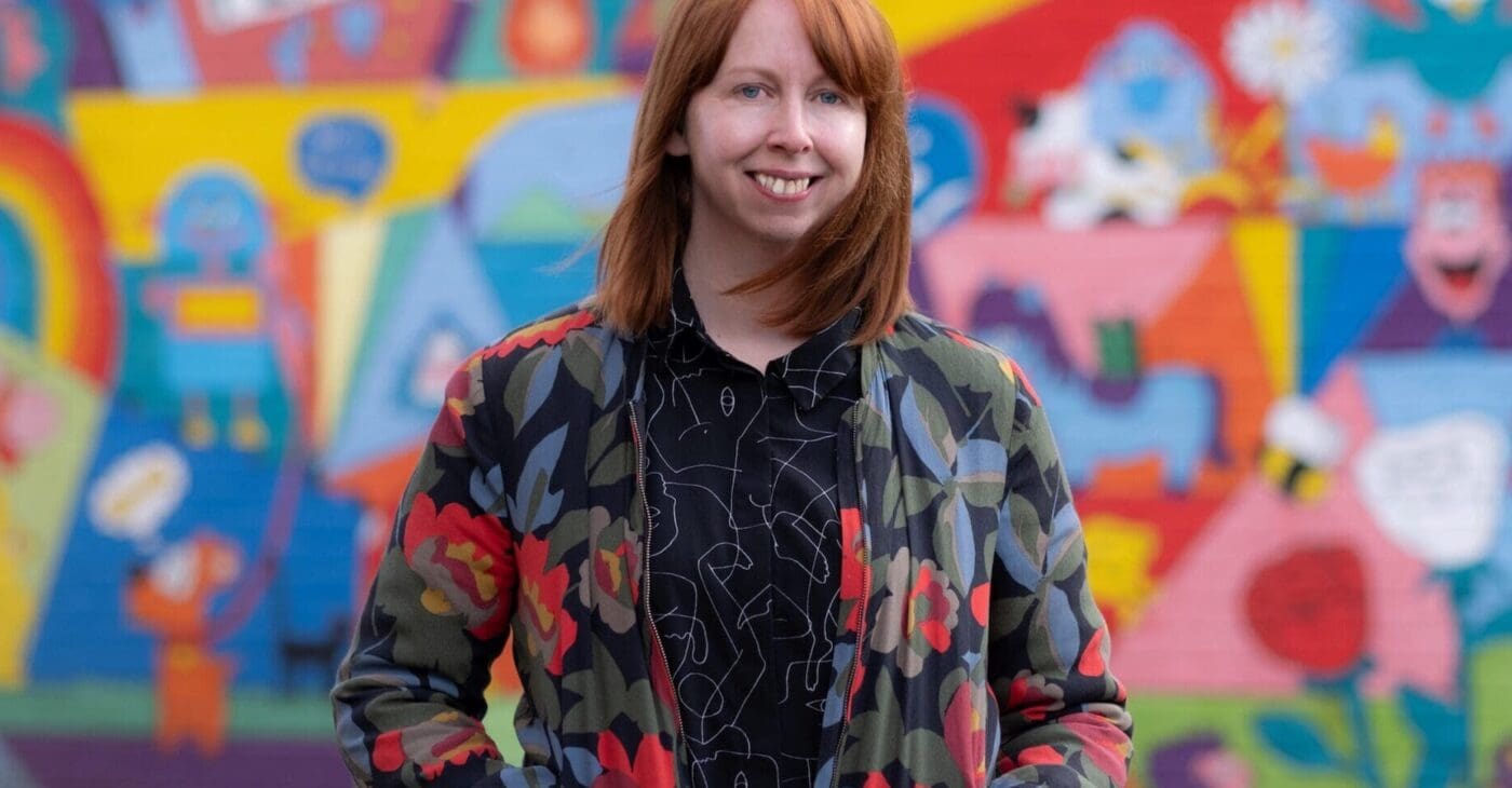 A white woman with ginger hair in her thirties wears a bomber jacket and stands in front of a colourful graffiti wall outside