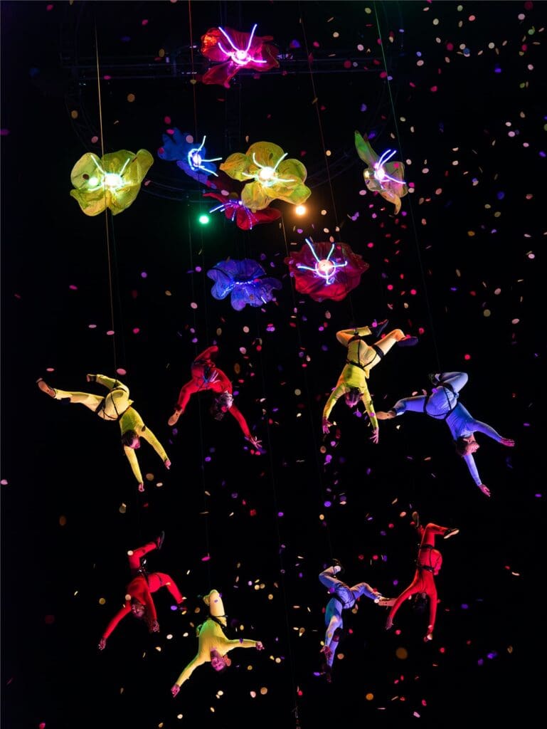 A group of acrobats and aerialists are dangling in the night sky from a huge flower-shaped structure with confetti all around them.