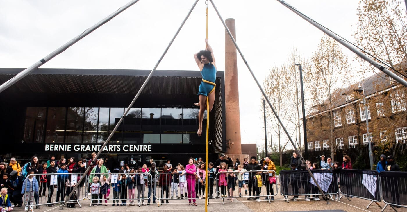 An aerialist holds herself up on a rope high in the sky in front of bernie grant arts centre