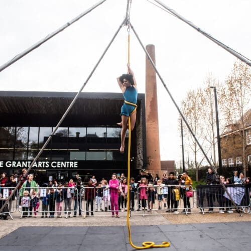 An aerialist holds herself up on a rope high in the sky in front of bernie grant arts centre