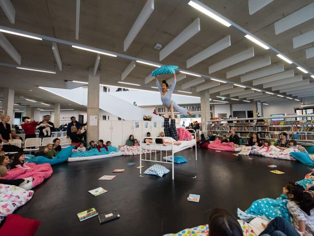 two acrobats perform a daring move in the middle of a library with children sat all around them