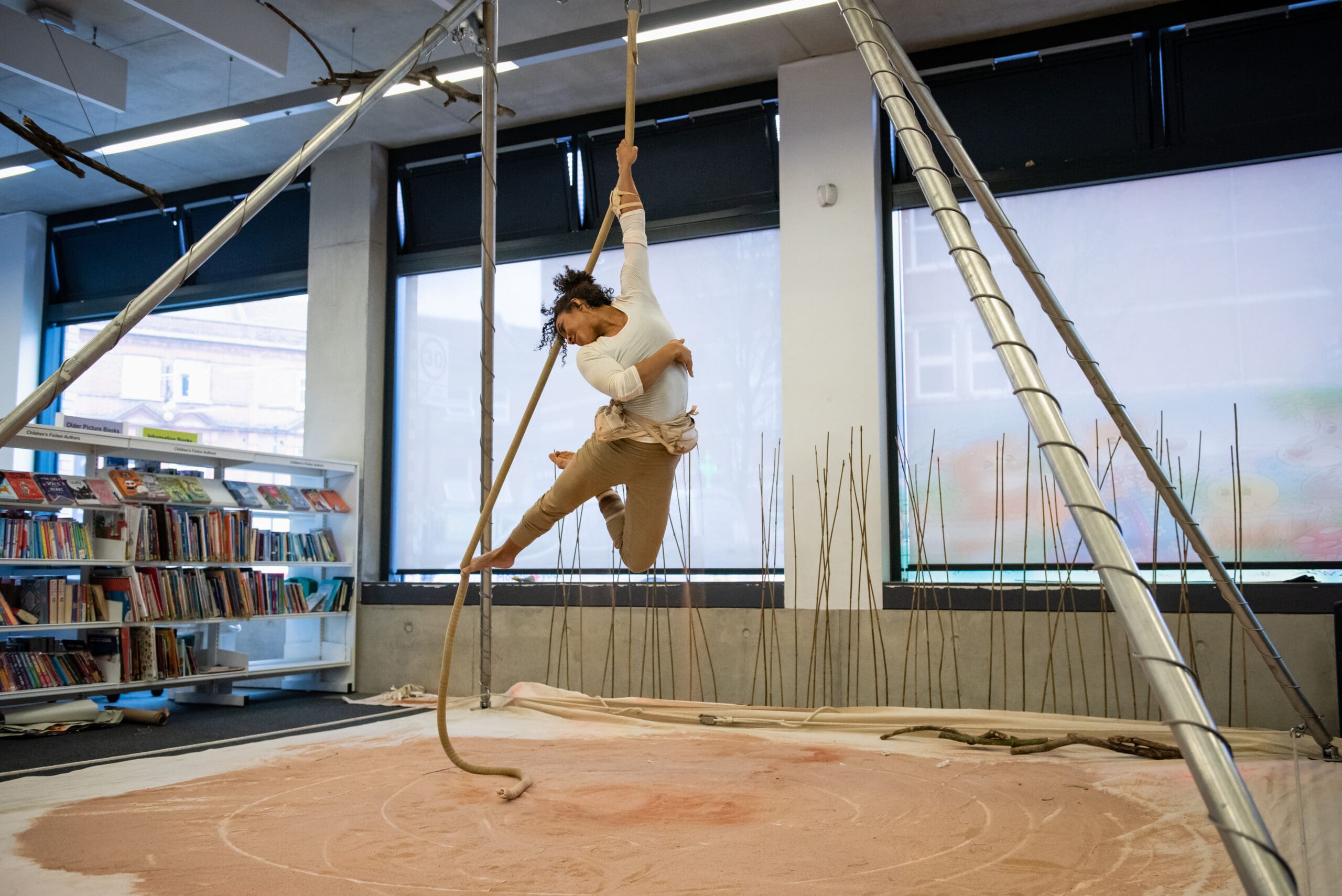 An aerialist pulls herself up on a rope attached to an aerial rig in the middle of a library surrounded by books