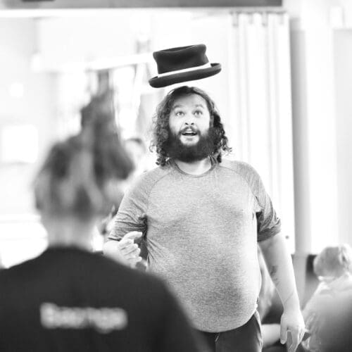 A black and white image of a Half Egyptian, Half Scottish gentleman with dark curly hair, and a full beard, gazes up at a felt top hat floating above his head.