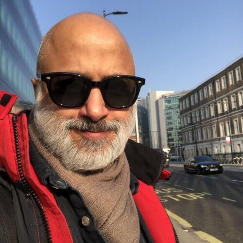 A British-Indian man with bald head and beard wearing sunglasses and a red jacket