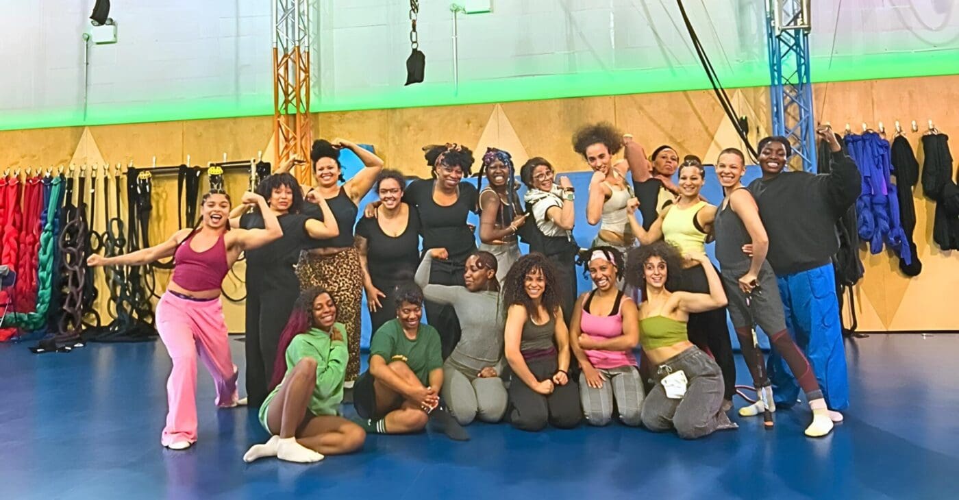 A group of 18 women and non-binary people from the Global Majority stand smiling together in a circus rehearsal space. They wear casual sports clothes and have a mixture of aerial ropes behind them.